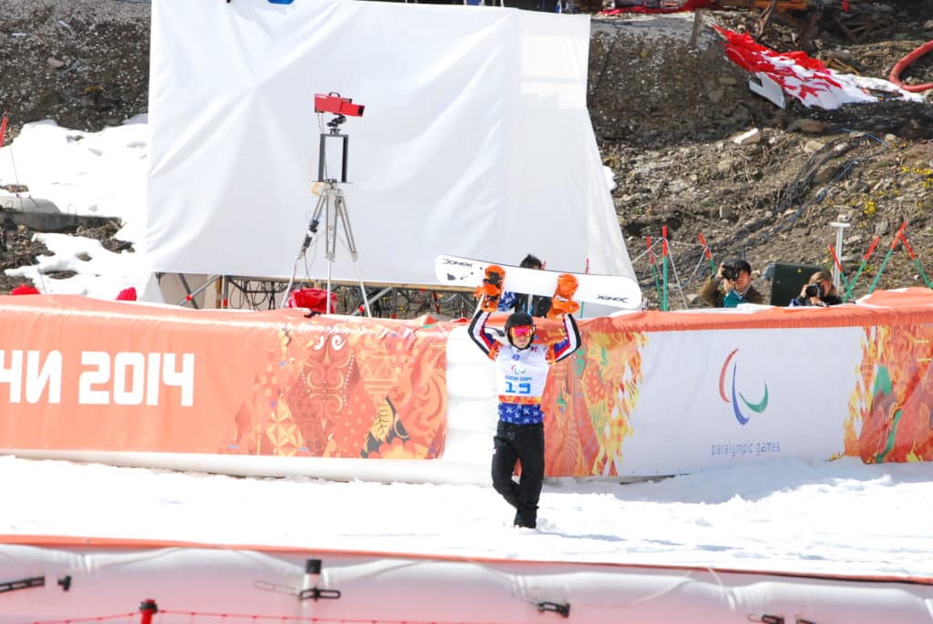 Paralympic winter snowboarder wearing bib number 15 raises both arms holding snowboard on the snow at a Sochi 2014 competition venue. 