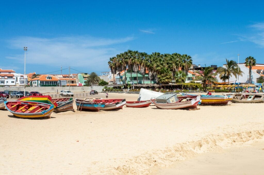 Colorful wooden fishing boats pulled onto a sandy beach in Cabo Verde, with palm trees and low-rise buildings behind them.